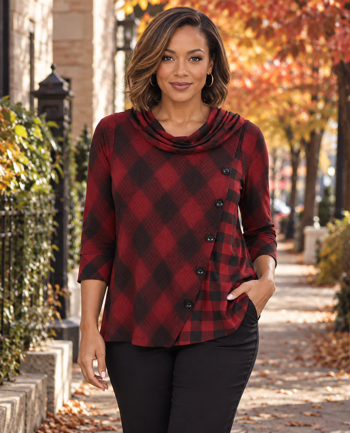 A woman stands on an outdoor sidewalk wearing a red and black buffalo plaid cowl neck top with black buttons and black pants.