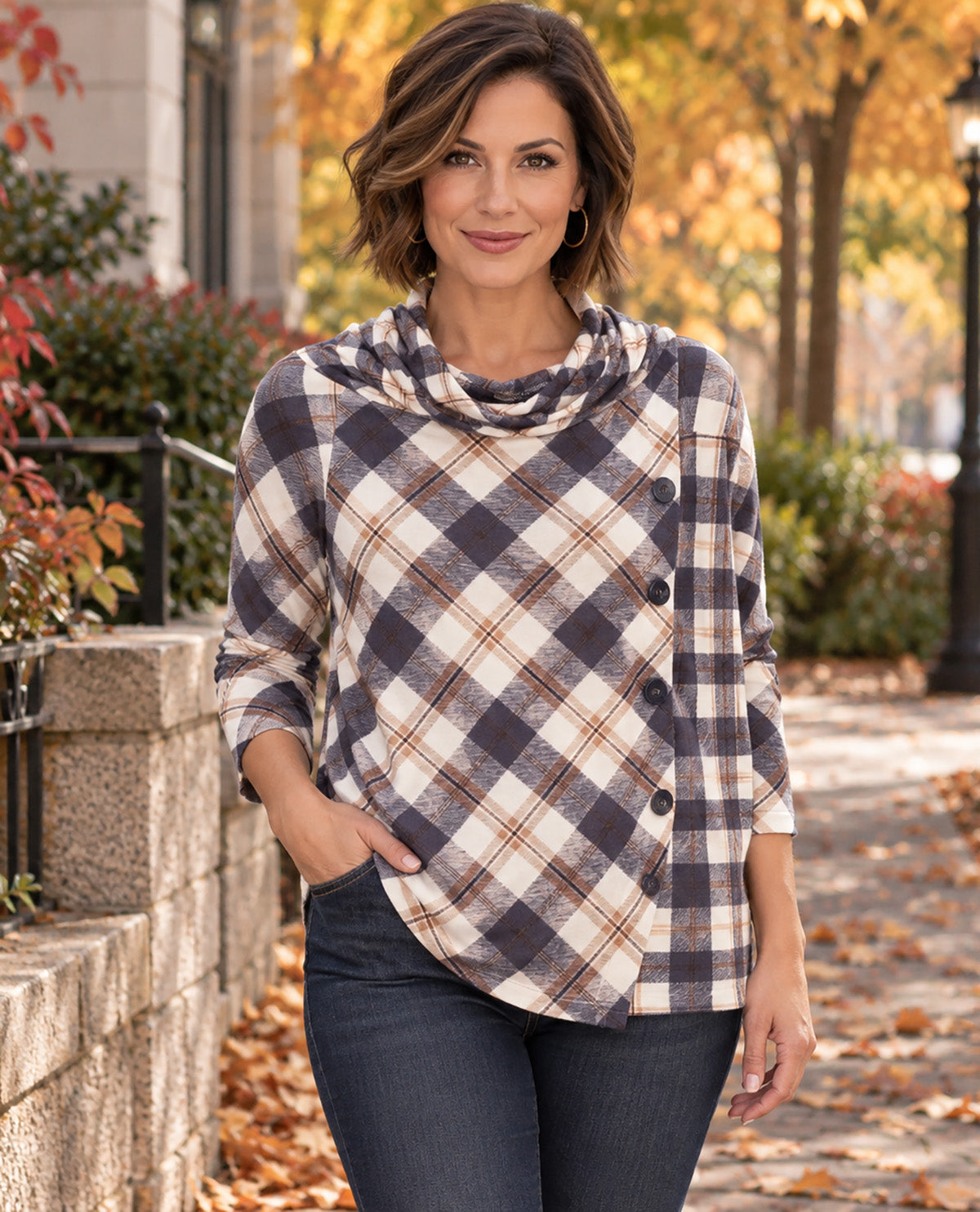 A woman wearing a plaid cowl-neck top with dark buttons, paired with dark jeans, standing outdoors on a path with autumn trees in the background.