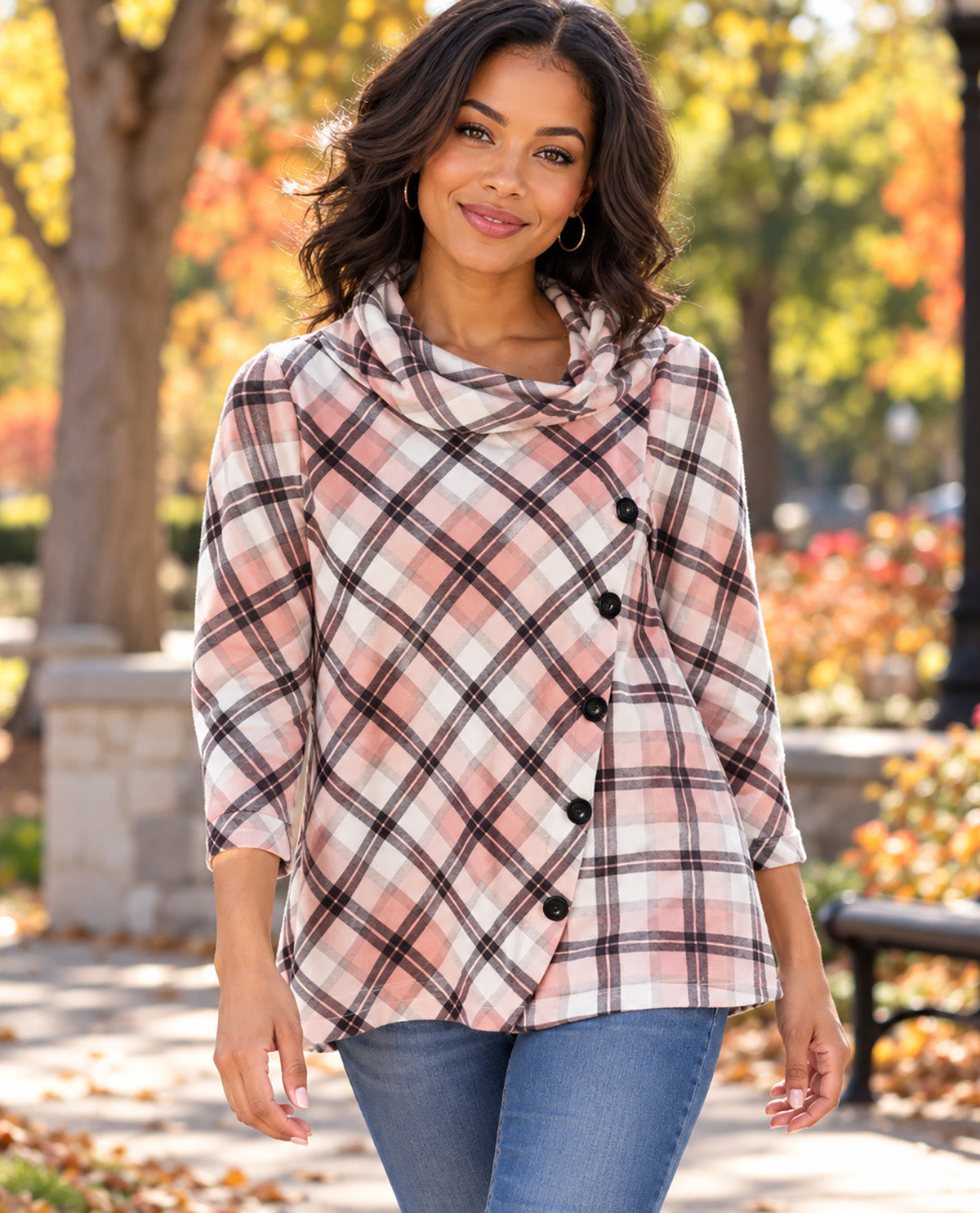 A woman wearing a pink, white, and black plaid wrap-style cowl-neck top with blue jeans, standing outdoors in a park with autumn trees in the background.