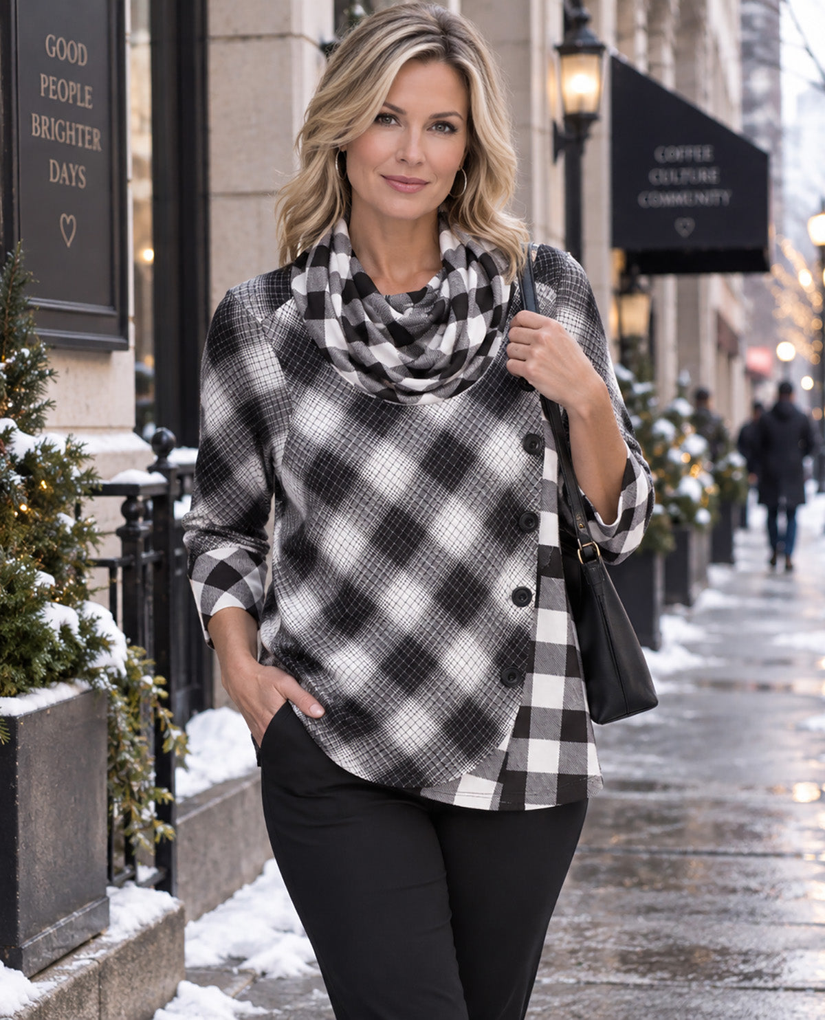A woman stands on a snowy city sidewalk wearing a black and white buffalo plaid wrap-style top with a matching cowl neck scarf, black pants, and carrying a black shoulder bag.