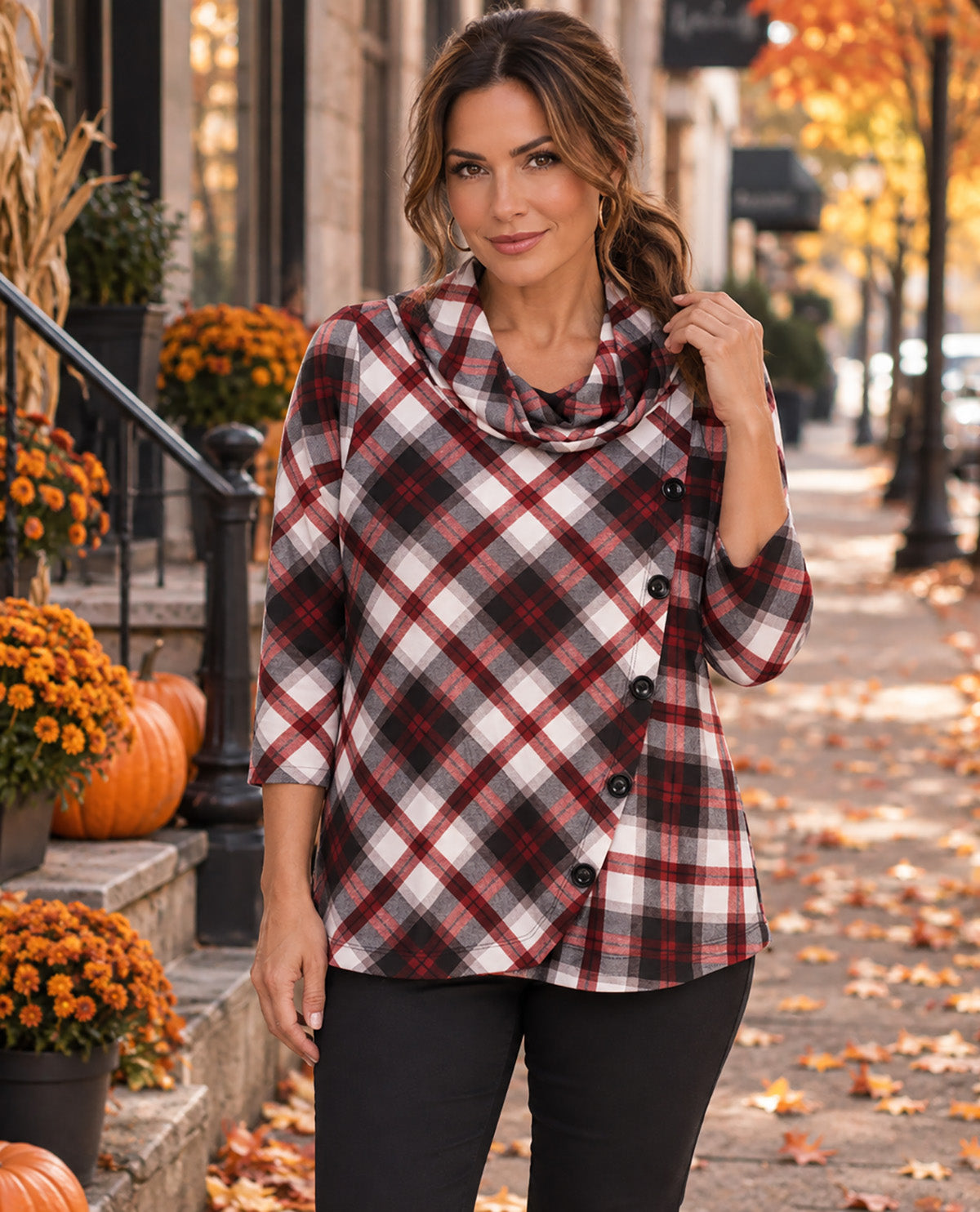 A woman stands on an autumn city sidewalk wearing a red, black, and white plaid wrap-style cowl-neck top with black pants. Pumpkins, orange flowers, and fallen leaves are visible in the background.