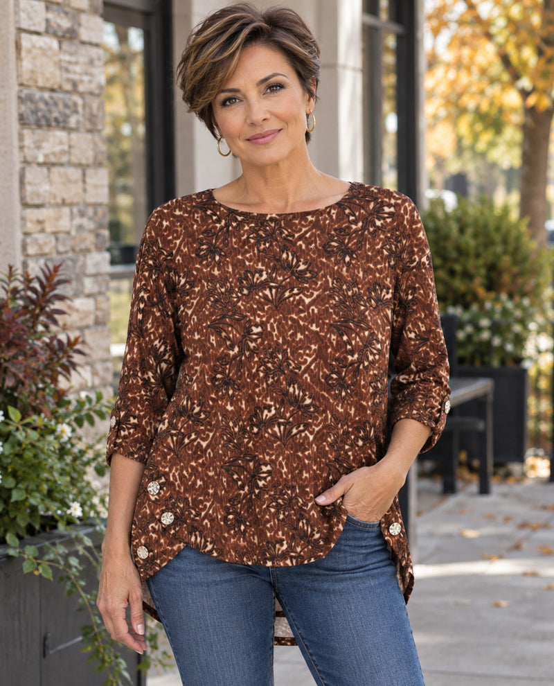A woman wearing a brown and beige abstract patterned top with three-quarter sleeves and blue jeans, standing outdoors in front of a building with stone walls and plants.