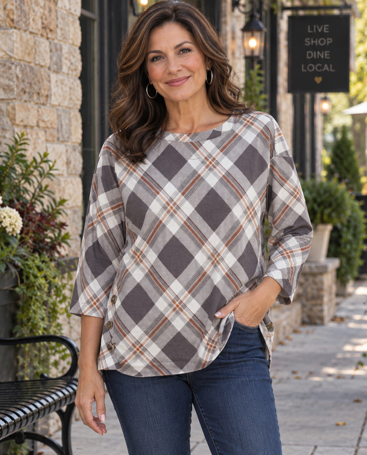 A woman wearing a gray, white, and brown plaid three-quarter sleeve top with blue jeans, standing outdoors on a sidewalk in front of a stone building.
