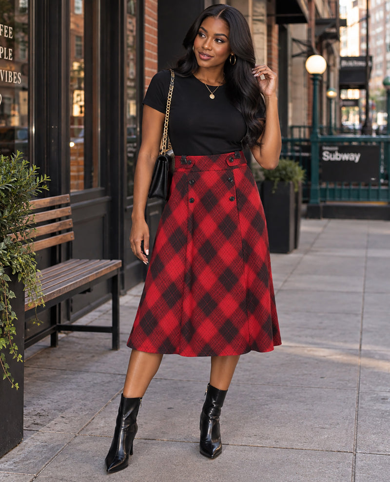 A woman stands on a city sidewalk wearing a black short-sleeve top and a red-and-black plaid high-waisted midi skirt, paired with black ankle boots and a small black shoulder bag.