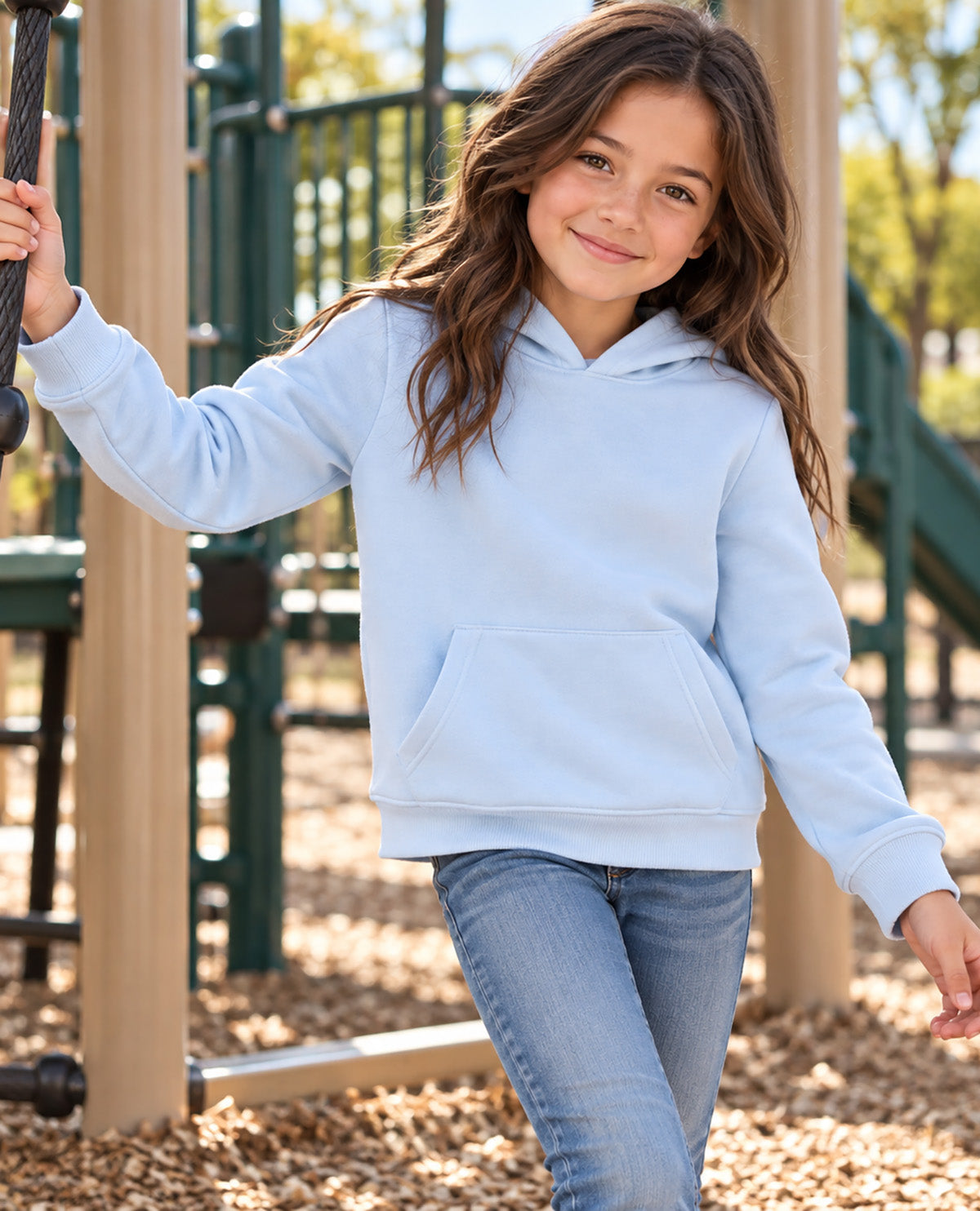 A young girl with long brown hair stands on a playground wearing a light blue pullover hoodie and blue jeans, smiling at the camera.