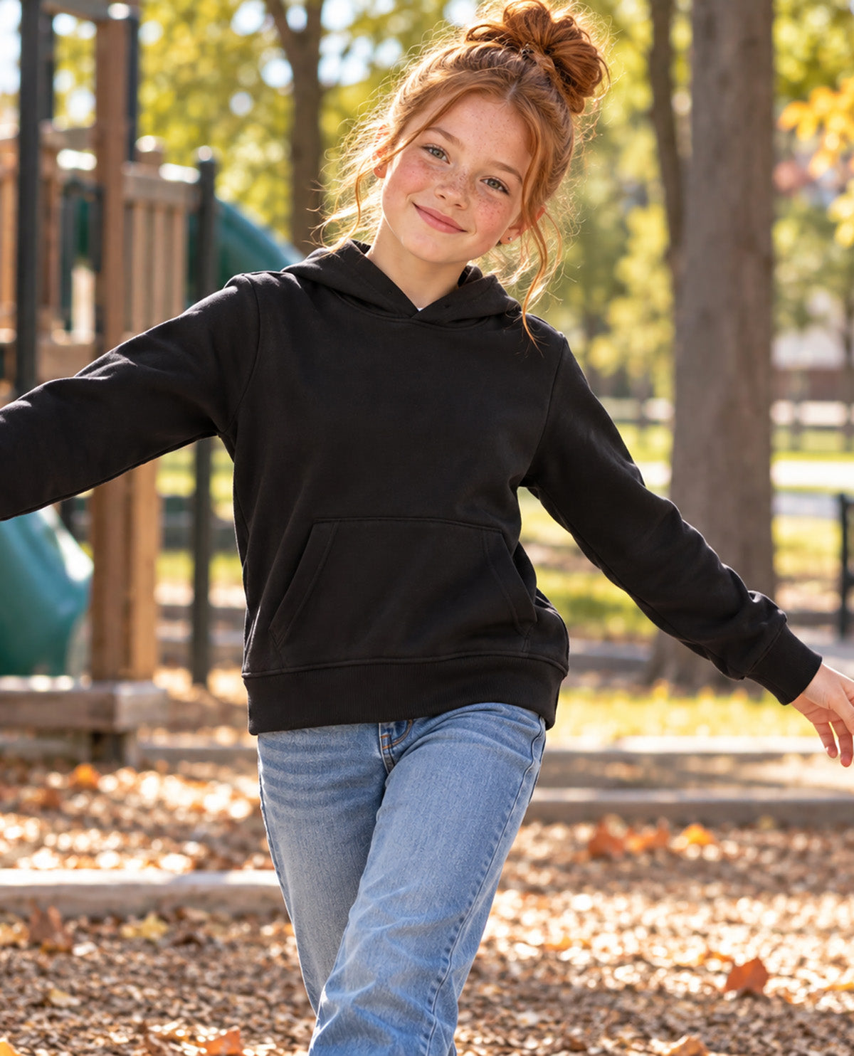 A young girl with red hair in a bun wearing a black pullover hoodie and light blue jeans, standing on a playground with fallen leaves in the background.