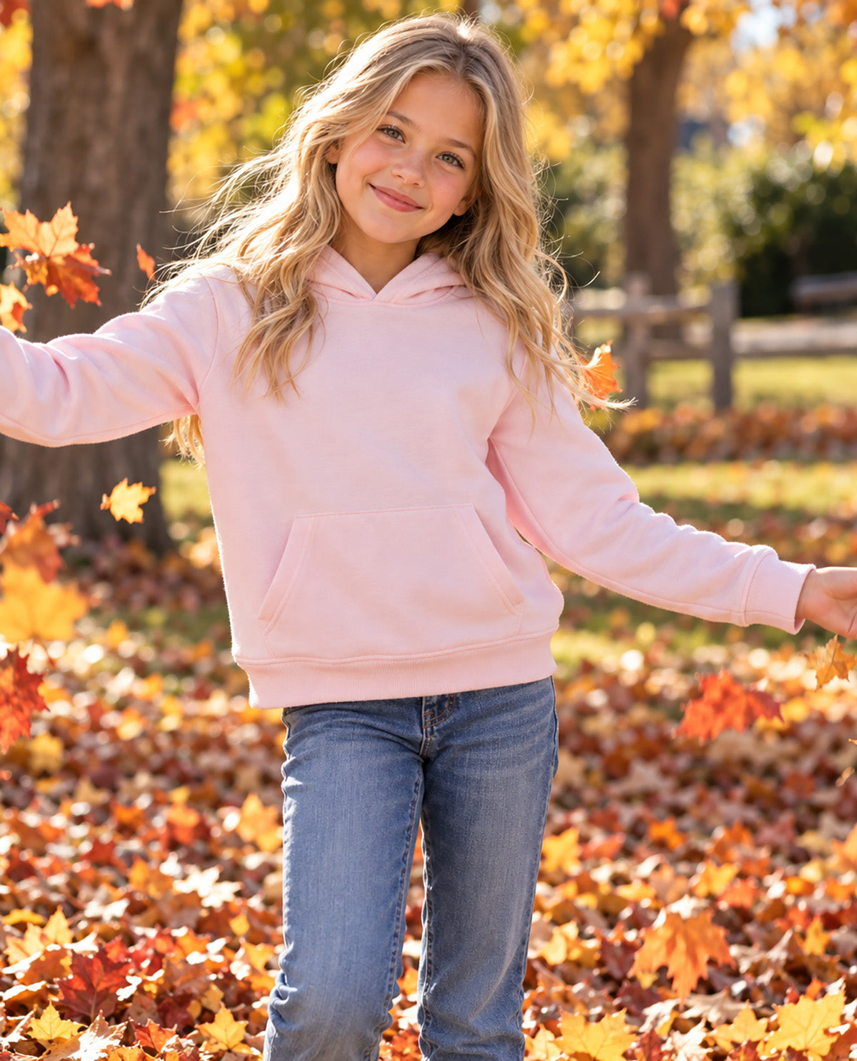 A young girl with blonde hair stands outdoors in a park covered with fallen autumn leaves, wearing a light pink pullover hoodie and blue jeans.