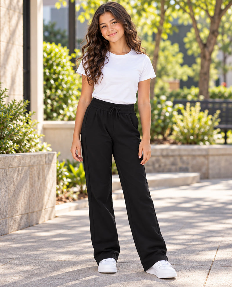 A young girl stands outdoors on a paved walkway wearing black wide-leg drawstring pants, a white short-sleeve T-shirt, and white sneakers.