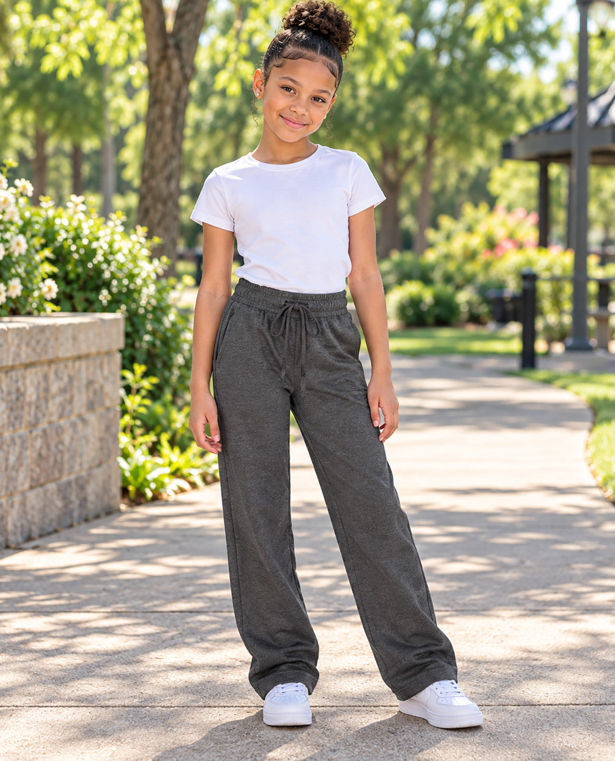 A young girl stands on a paved path in a sunny park, wearing dark gray wide-leg drawstring sweatpants, a white t-shirt, and white sneakers.