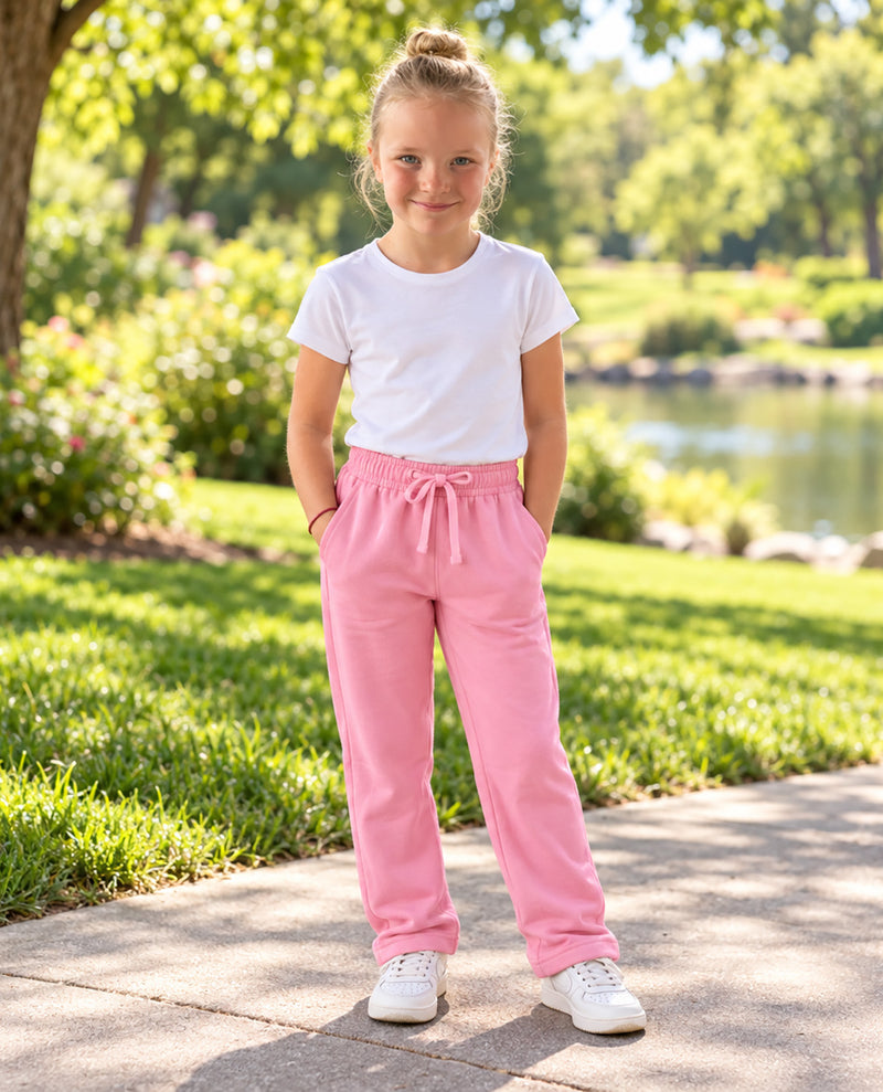 A young girl stands on a paved path in a sunny park, wearing pink drawstring sweatpants, a white t-shirt, and white sneakers.