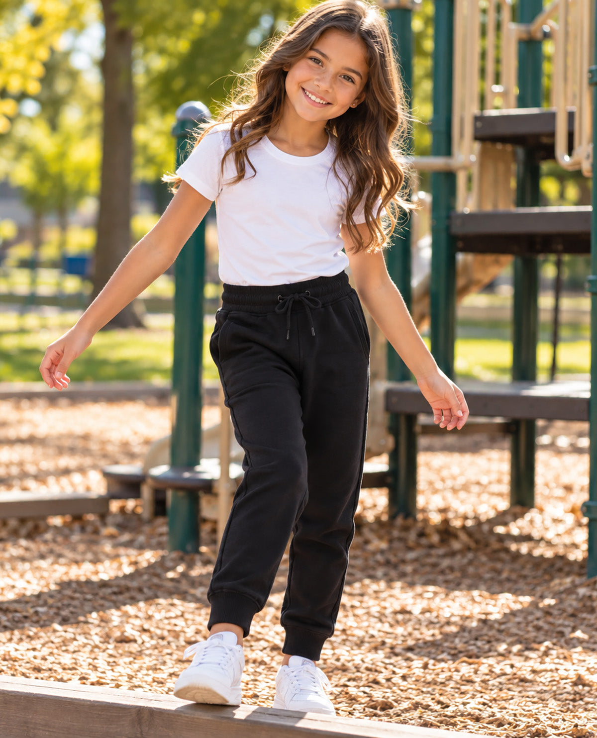 A young girl smiling while walking on a playground, wearing a white short-sleeve t-shirt, black jogger sweatpants, and white sneakers.