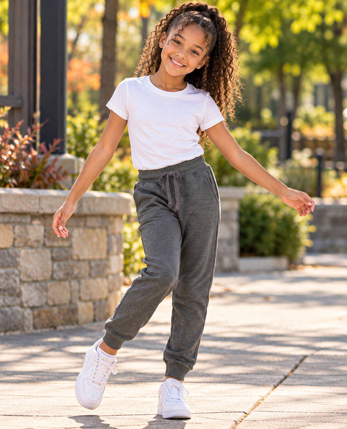 A young girl with curly hair is smiling and posing outdoors on a paved walkway. She is wearing a white short-sleeve t-shirt, dark gray jogger sweatpants with a drawstring waist, and white sneakers.