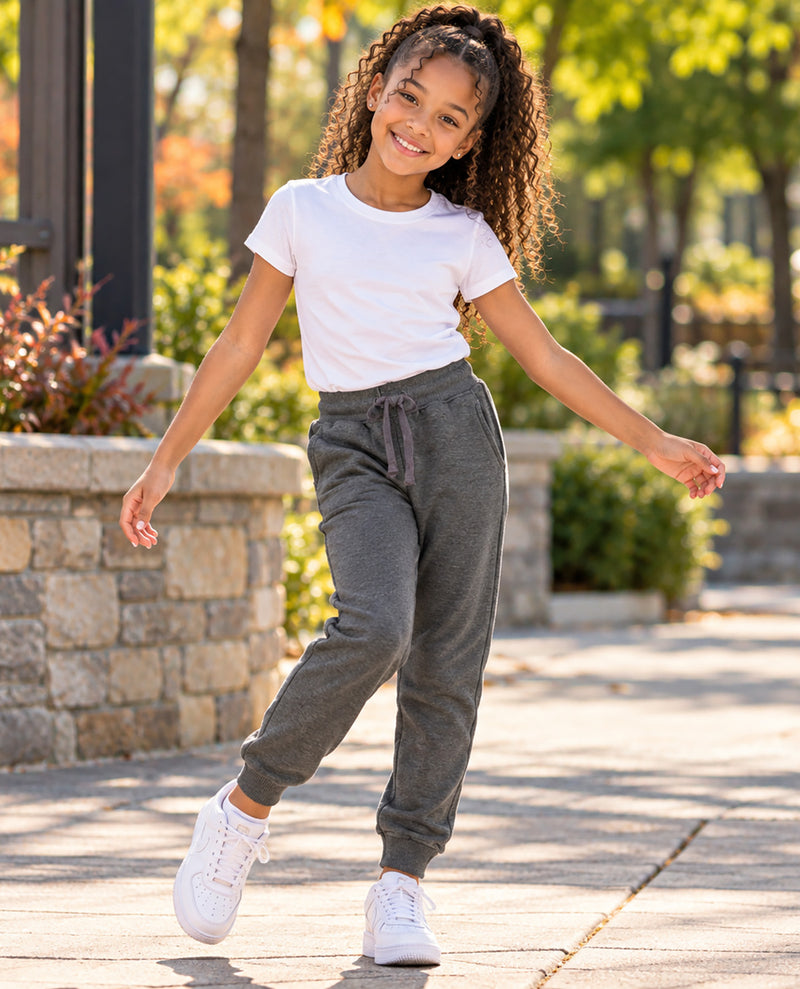 A young girl with curly hair is smiling and posing outdoors on a paved walkway. She is wearing a white short-sleeve t-shirt, dark gray jogger sweatpants with a drawstring waist, and white sneakers.