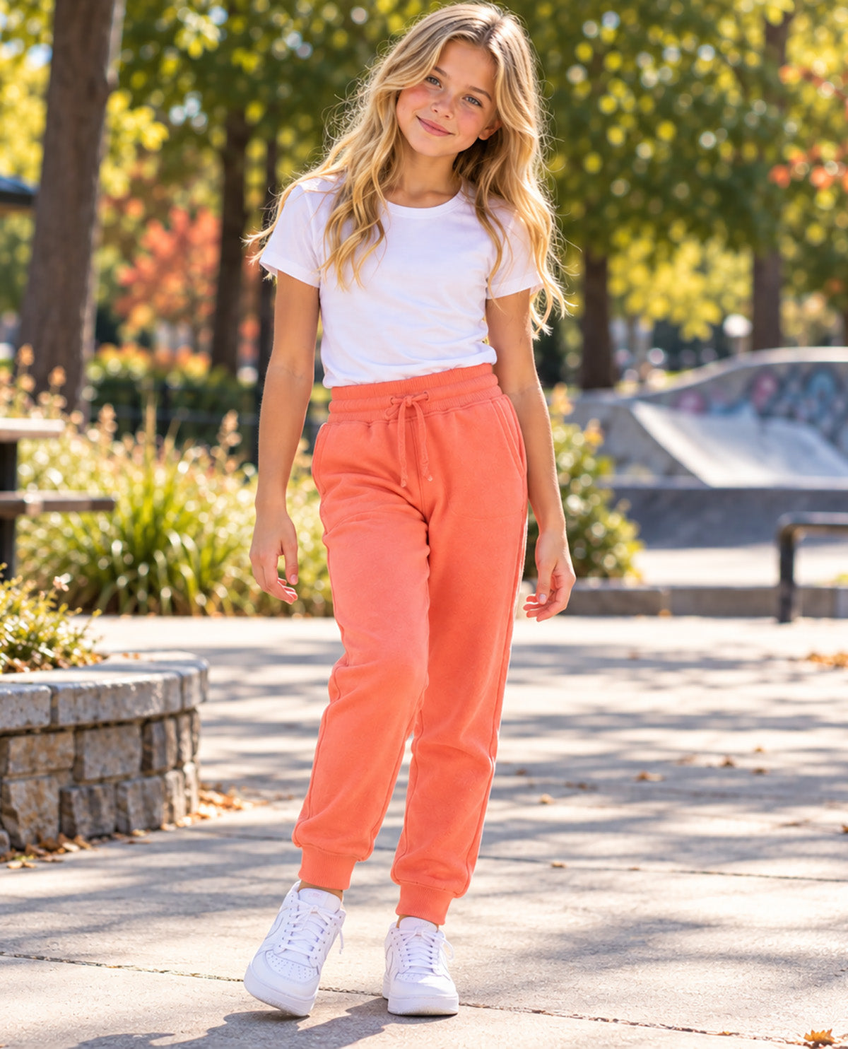 A young girl wearing a white t-shirt and bright coral orange sweatpants with white sneakers, standing on a paved path in an outdoor park setting with trees and a skatepark in the background.