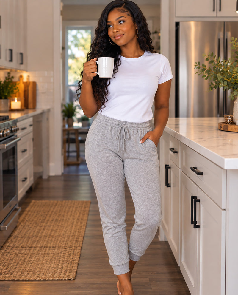 A woman stands in a bright, modern kitchen wearing a white crewneck t-shirt and light gray heather jogger sweatpants, holding a white mug.