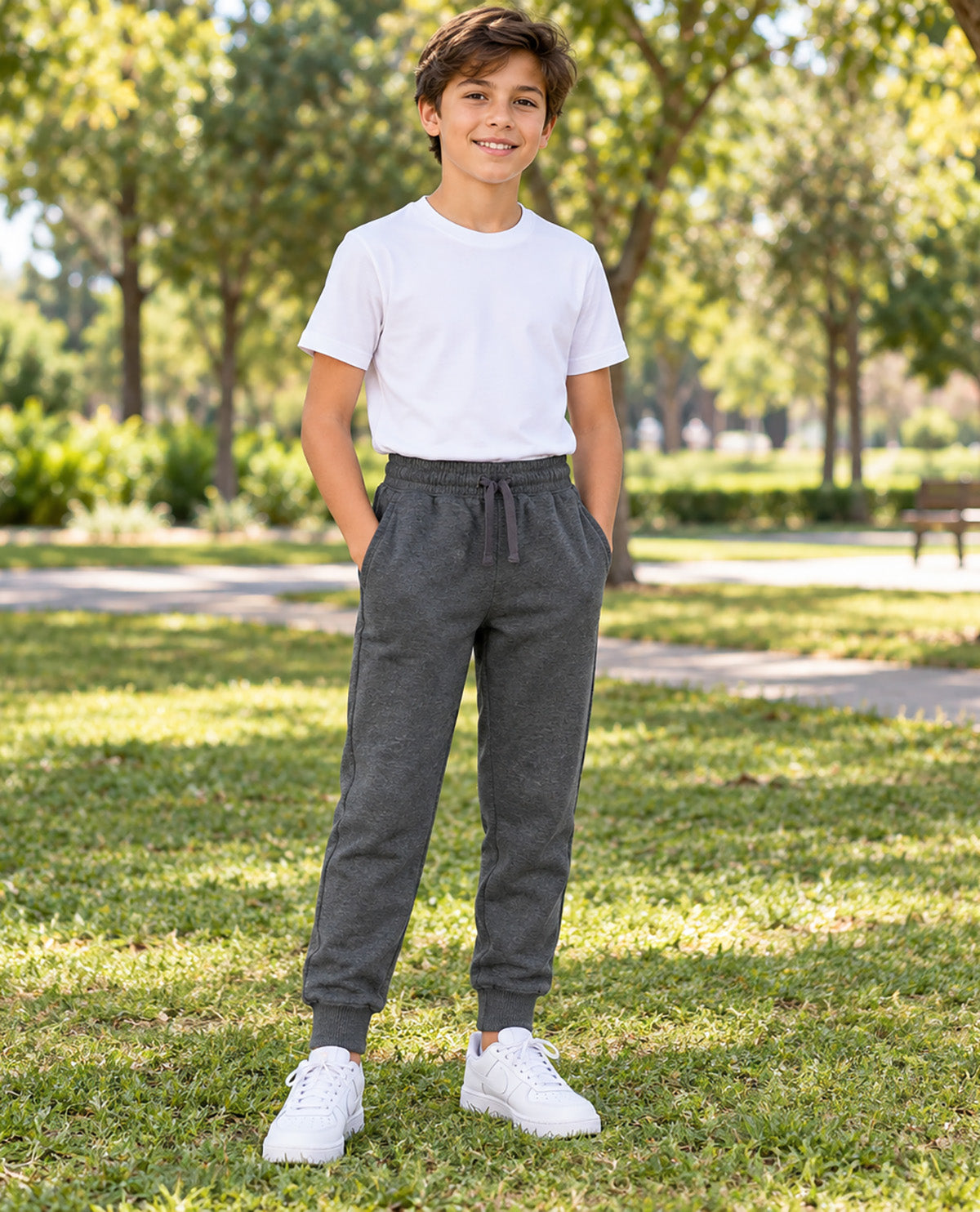 A young boy standing in a sunny park wearing dark gray jogger sweatpants, a white t-shirt, and white sneakers.