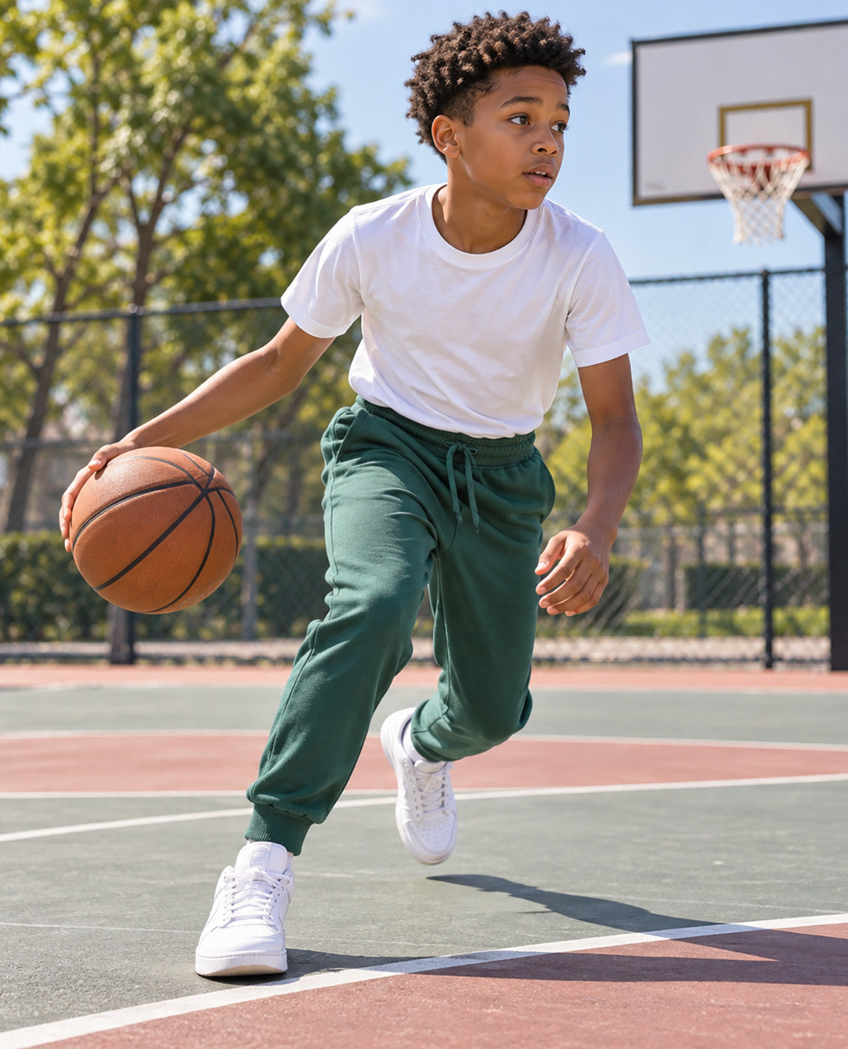 A young boy dribbling a basketball on an outdoor court, wearing a white t-shirt, dark green jogger sweatpants, and white sneakers.