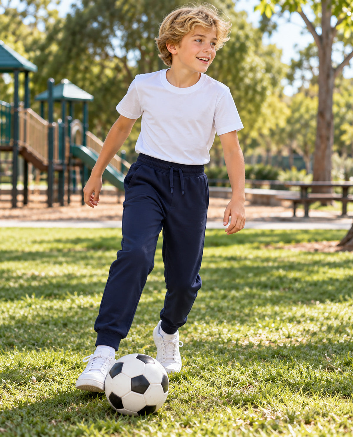 A young boy wearing a white t-shirt and navy blue sweatpants is playing with a soccer ball on a grassy park lawn.