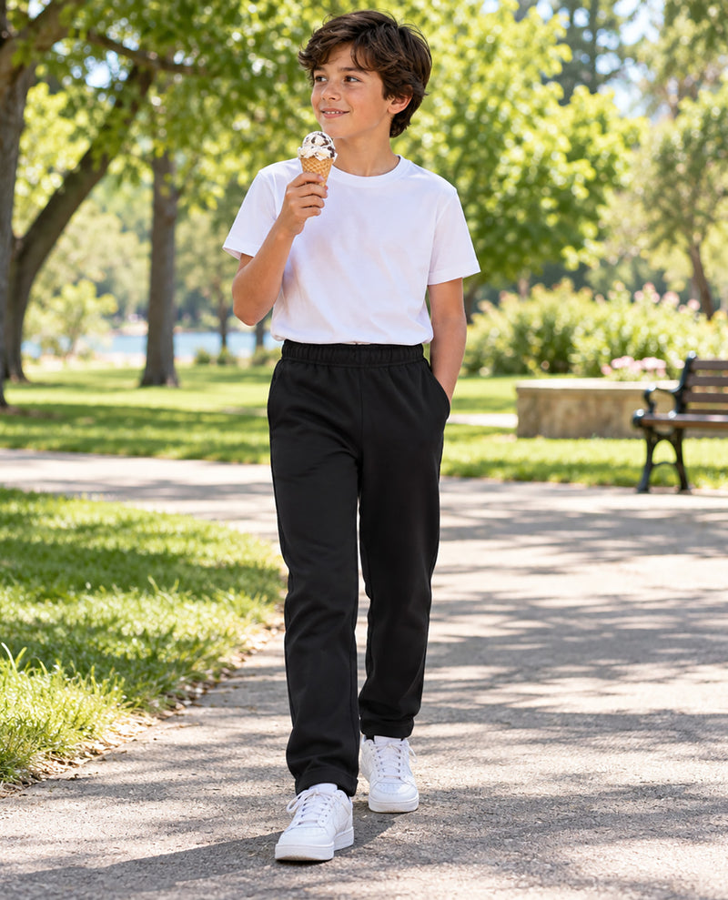 A boy walking on a sunny park path wearing a white crewneck t-shirt, black sweatpants, and white sneakers while holding an ice cream cone.