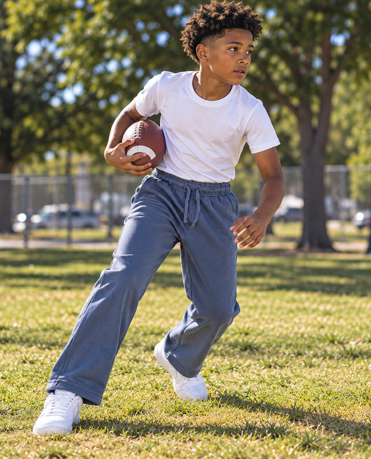 A boy running on a grassy field holding a football, wearing a white t-shirt, blue sweatpants, and white sneakers.