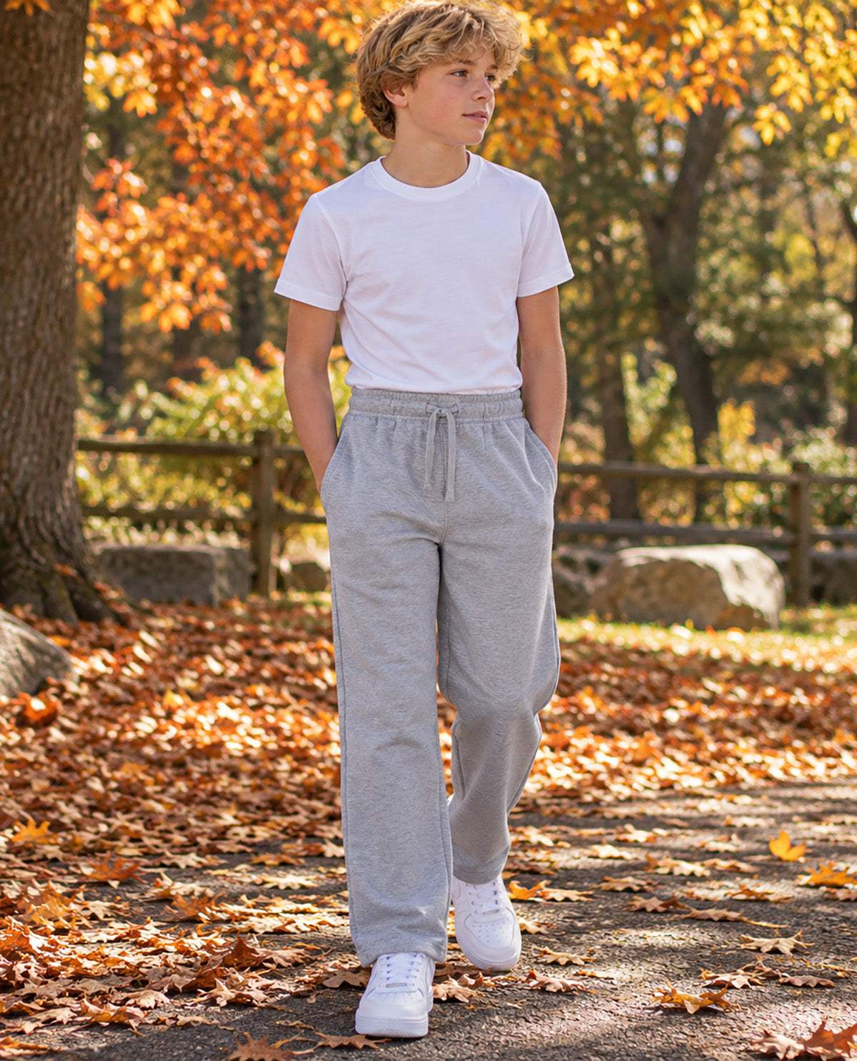 A young boy standing on an autumn leaf-covered path wearing a white crewneck t-shirt and light gray sweatpants.