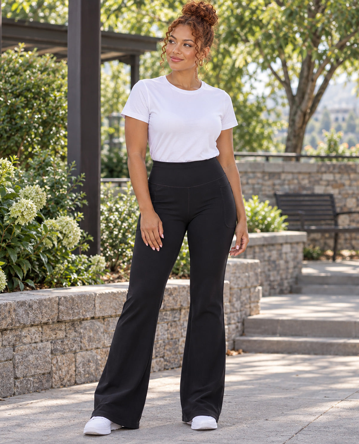 A woman stands outdoors on a paved walkway wearing black high-waisted flare pants, a white short-sleeve crewneck t-shirt, and white sneakers. The background features stone walls, green bushes, and trees.