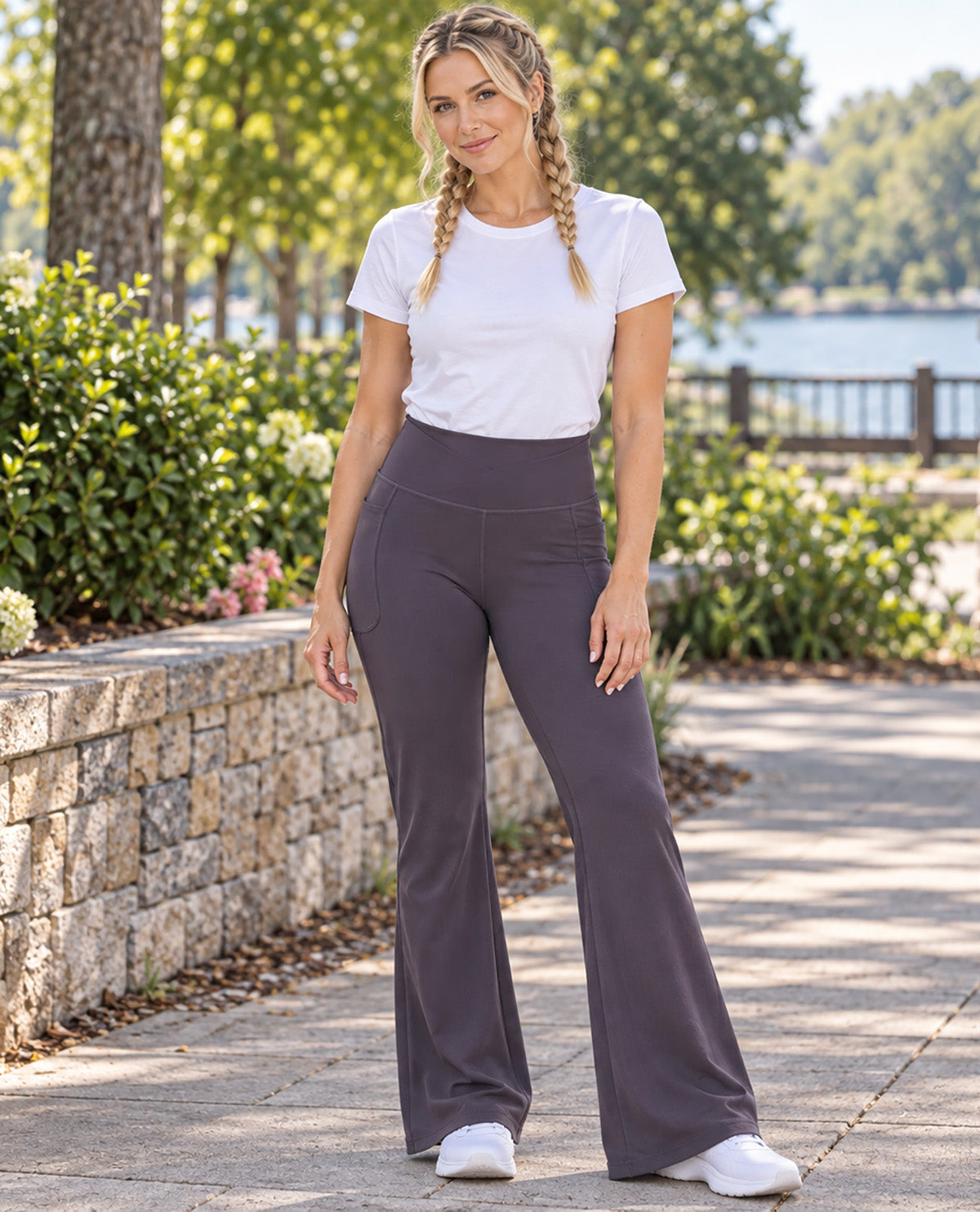 A woman stands outdoors on a paved walkway wearing high-waisted, wide-leg dark gray pants, a white short-sleeve crewneck t-shirt, and white sneakers. The background features a stone wall, green bushes, trees, and a body of water.