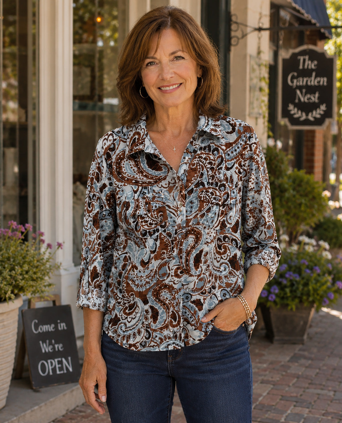 A woman stands on a brick sidewalk in front of a storefront wearing a long-sleeve button-up blouse with a brown, beige, and blue paisley pattern, paired with dark blue jeans.