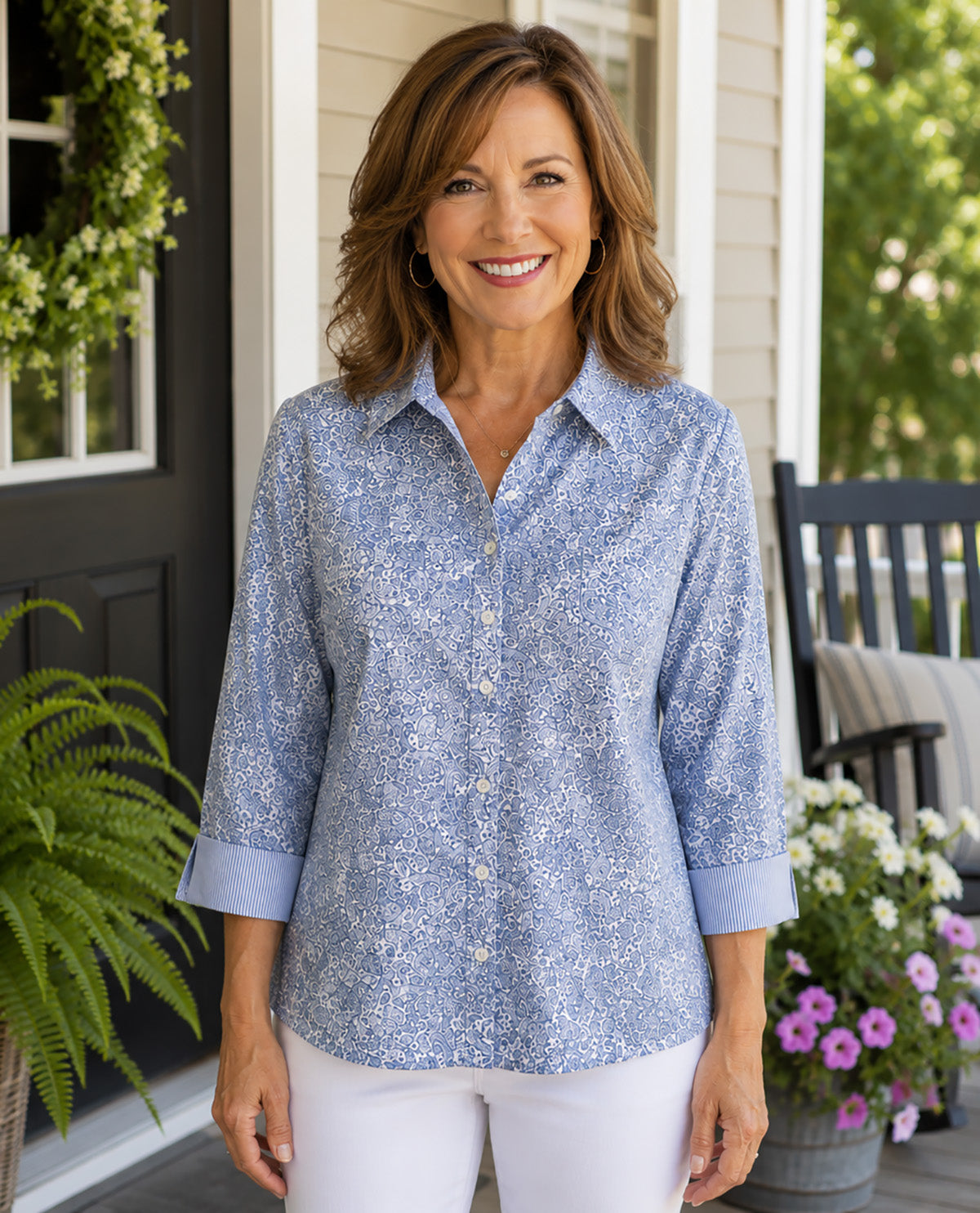 A smiling woman stands on a front porch wearing a light blue and white floral print button-down shirt with three-quarter sleeves and white pants.