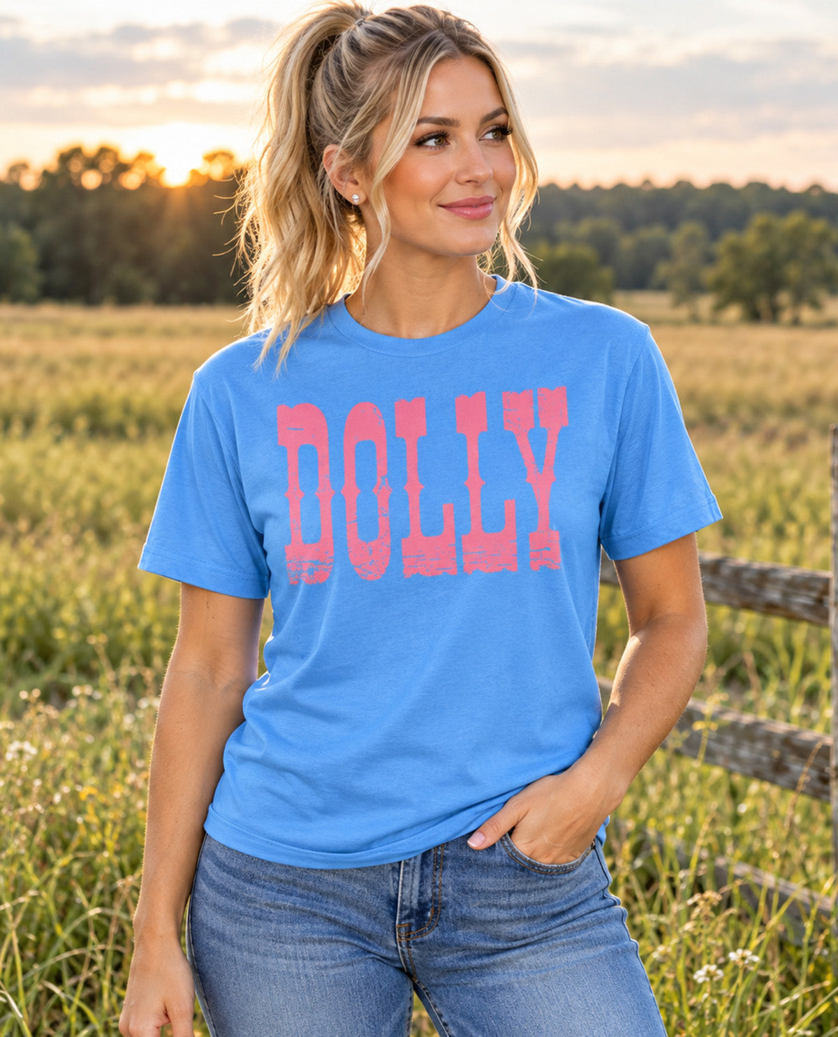 A woman wearing a bright blue t-shirt with 'DOLLY' printed in pink block letters, standing in a grassy field at sunset.