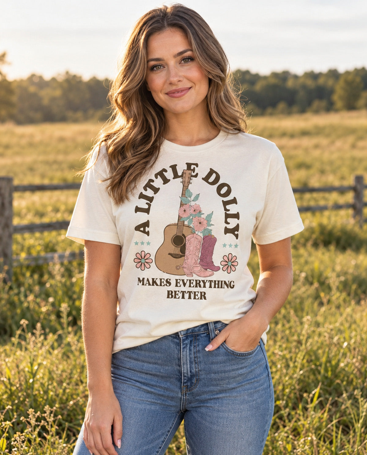 A woman standing in a grassy field at sunset wearing a cream-colored graphic t-shirt with a western-themed design and blue jeans.