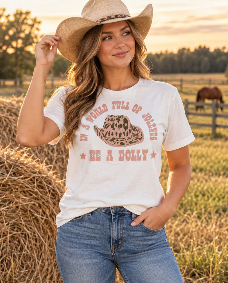 A woman wearing a white graphic t-shirt with a brown cowboy hat and blue jeans, standing in a sunny field with hay bales and a horse in the background. The t-shirt features a brown graphic of a cowboy hat and the text 'IN A WORLD FULL OF JOLENES BE A DOLLY'.