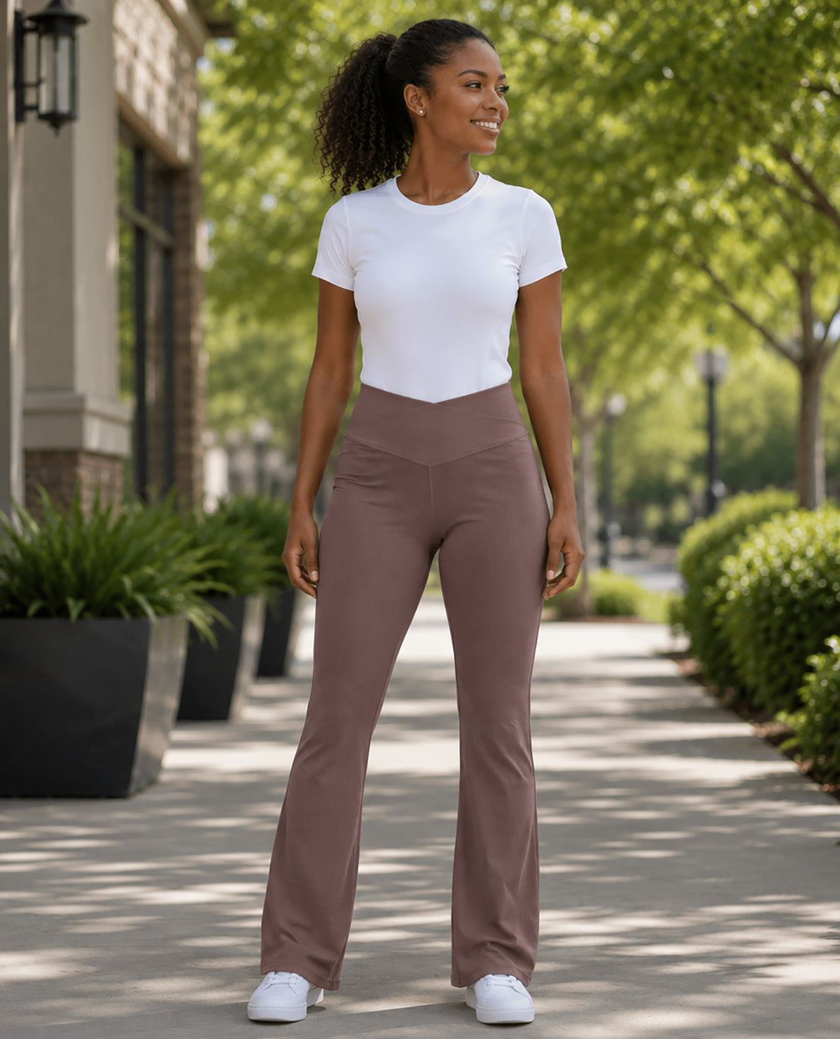 A woman stands on a tree-lined sidewalk wearing a fitted white short-sleeve top and high-waisted, straight-leg taupe pants with white sneakers.