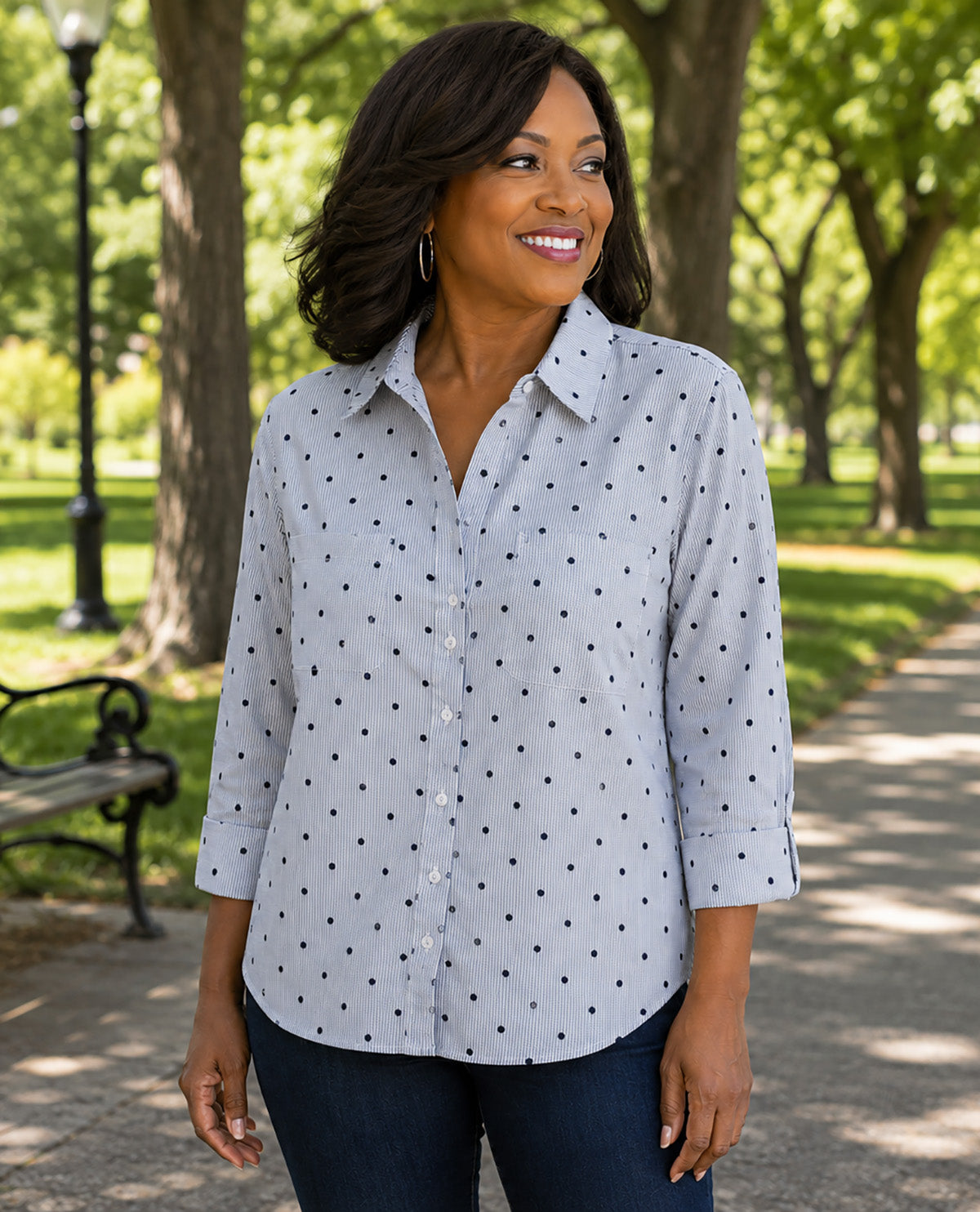 A woman wearing a light gray button-up shirt with a black polka dot pattern, standing in a sunny park with trees and a path in the background.