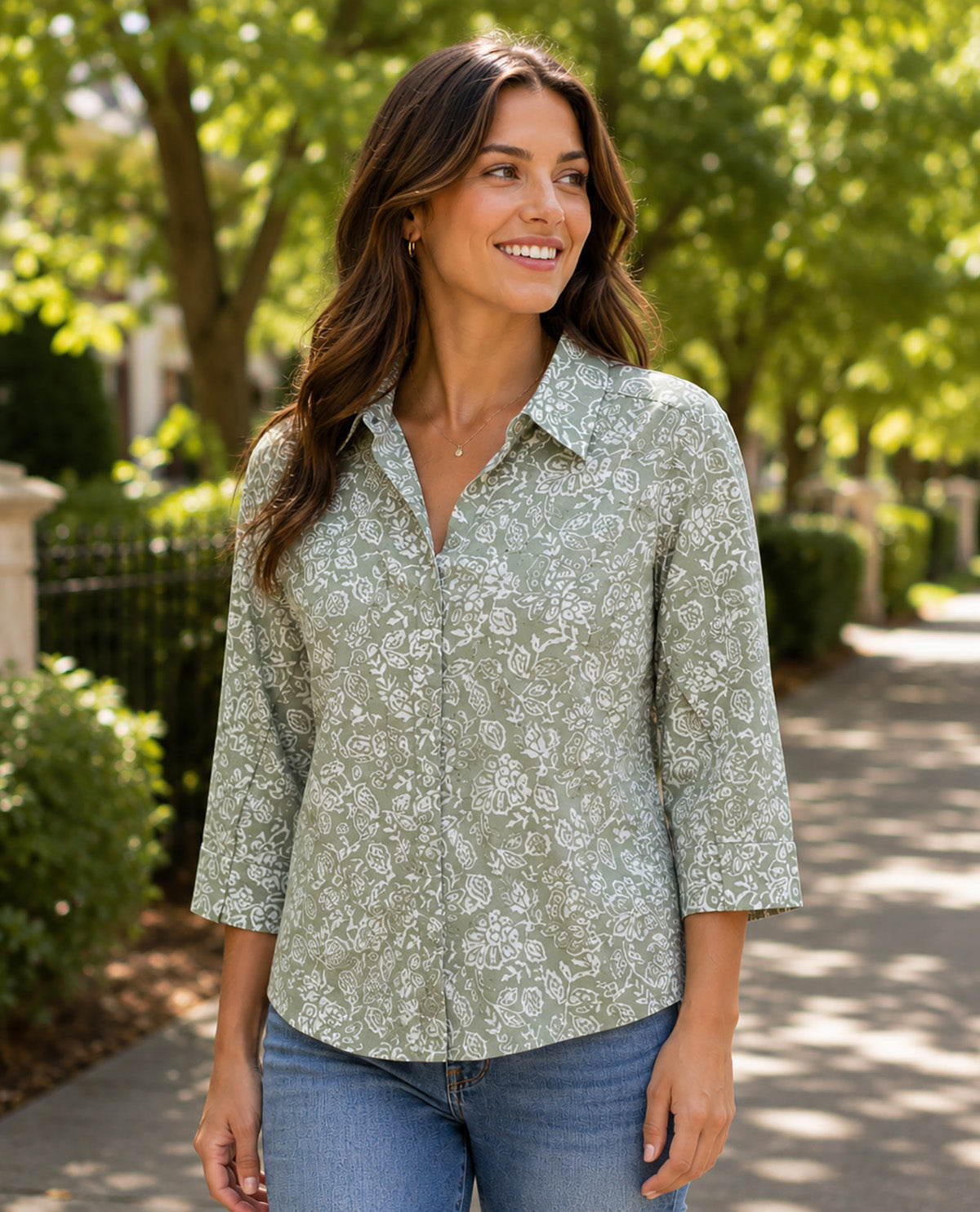 A woman wearing a sage green and white floral print button-down shirt with three-quarter sleeves, standing on a sunny outdoor path.
