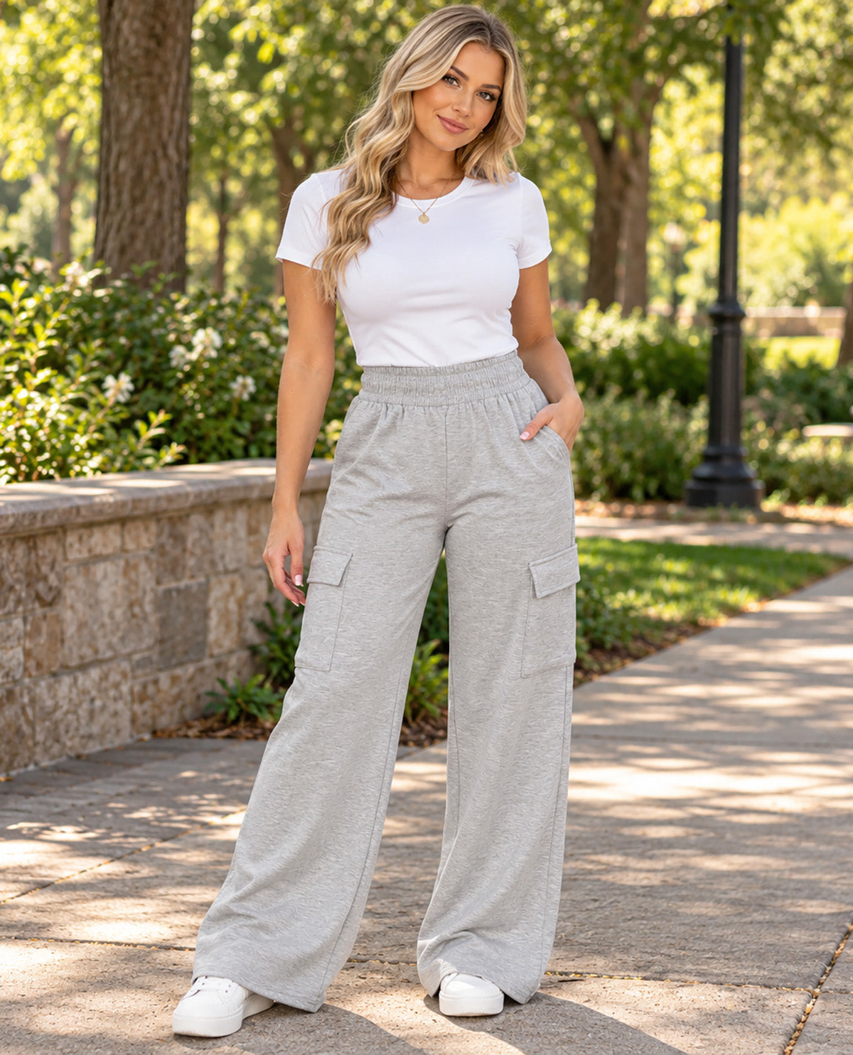 A woman modeling light gray wide-leg cargo pants with a white t-shirt and white sneakers, standing on a paved path in a sunny park with trees and greenery in the background.