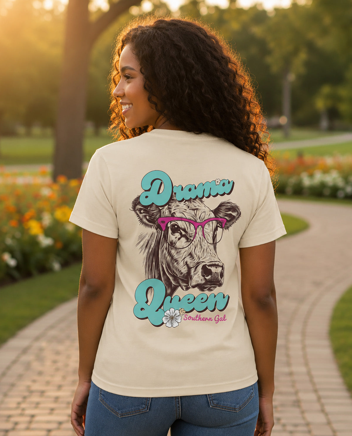 A woman standing on a paved path in a park, viewed from behind, wearing a cream-colored graphic t-shirt with a cow illustration and the words 'Drama Queen' in teal and pink.
