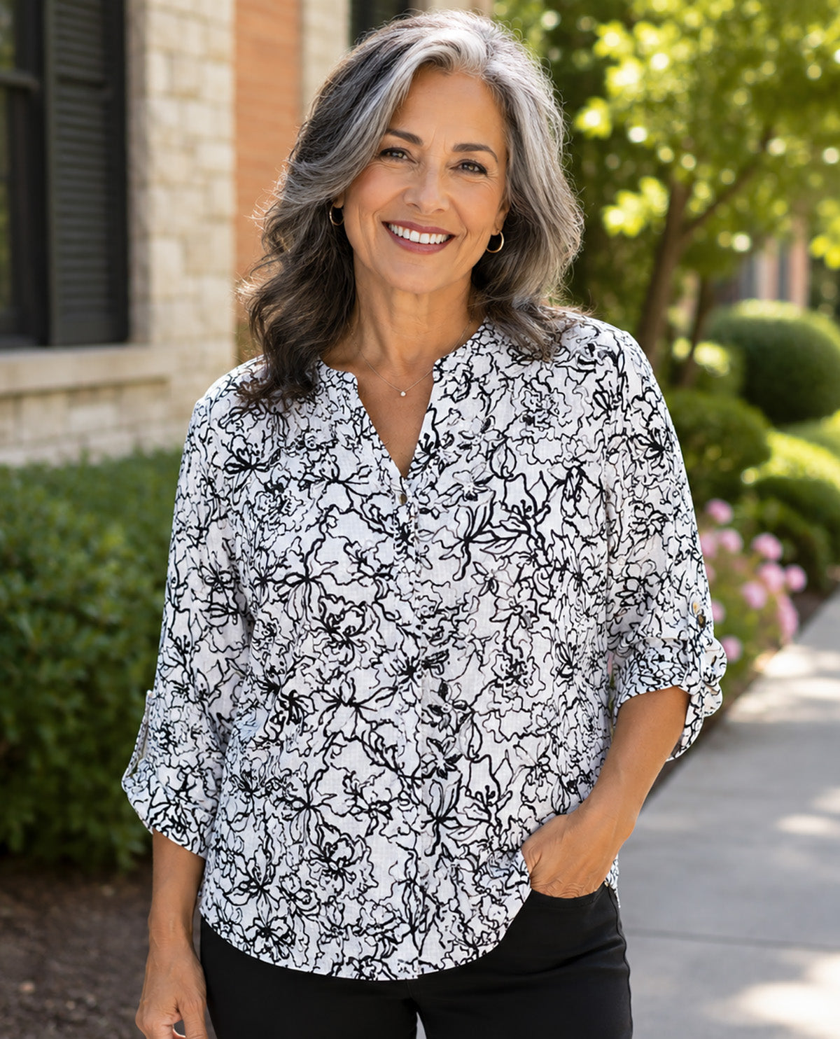 A smiling woman with gray hair stands outdoors wearing a black and white floral print blouse with three-quarter sleeves and black pants.