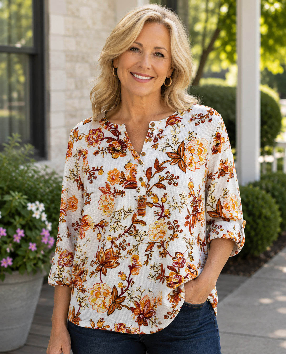 A smiling blonde woman wearing a white three-quarter sleeve blouse with an orange, brown, and yellow floral print, paired with dark blue jeans, standing outdoors in front of a house.