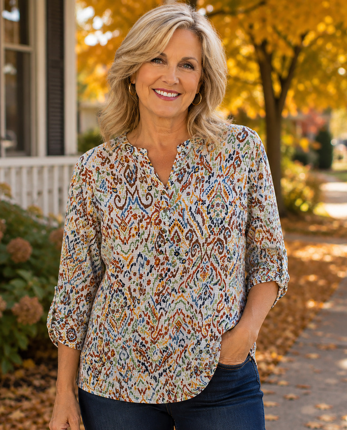 A smiling blonde woman wearing a patterned three-quarter sleeve blouse and dark blue jeans, standing outdoors on a path with autumn trees and a house in the background.