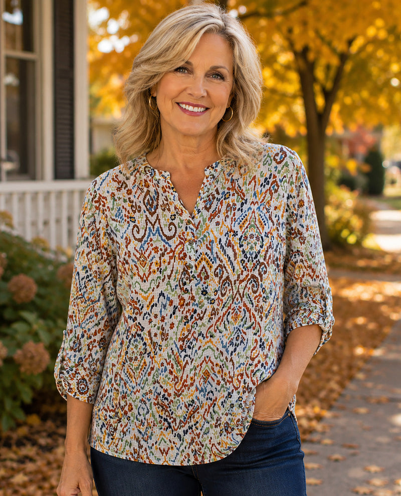 A smiling blonde woman wearing a patterned three-quarter sleeve blouse and dark blue jeans, standing outdoors on a path with autumn trees and a house in the background.
