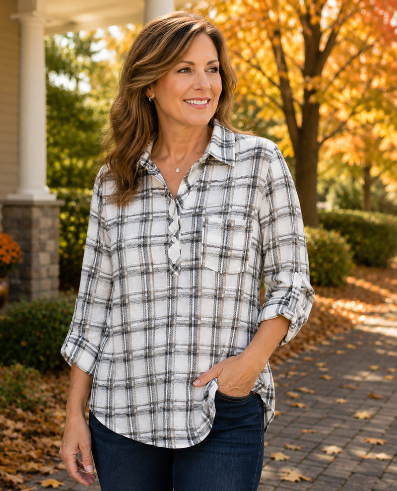 A woman wearing a white long-sleeve plaid button-up shirt with dark gray and black lines, paired with dark blue jeans, standing outdoors on a brick path with autumn trees in the background.