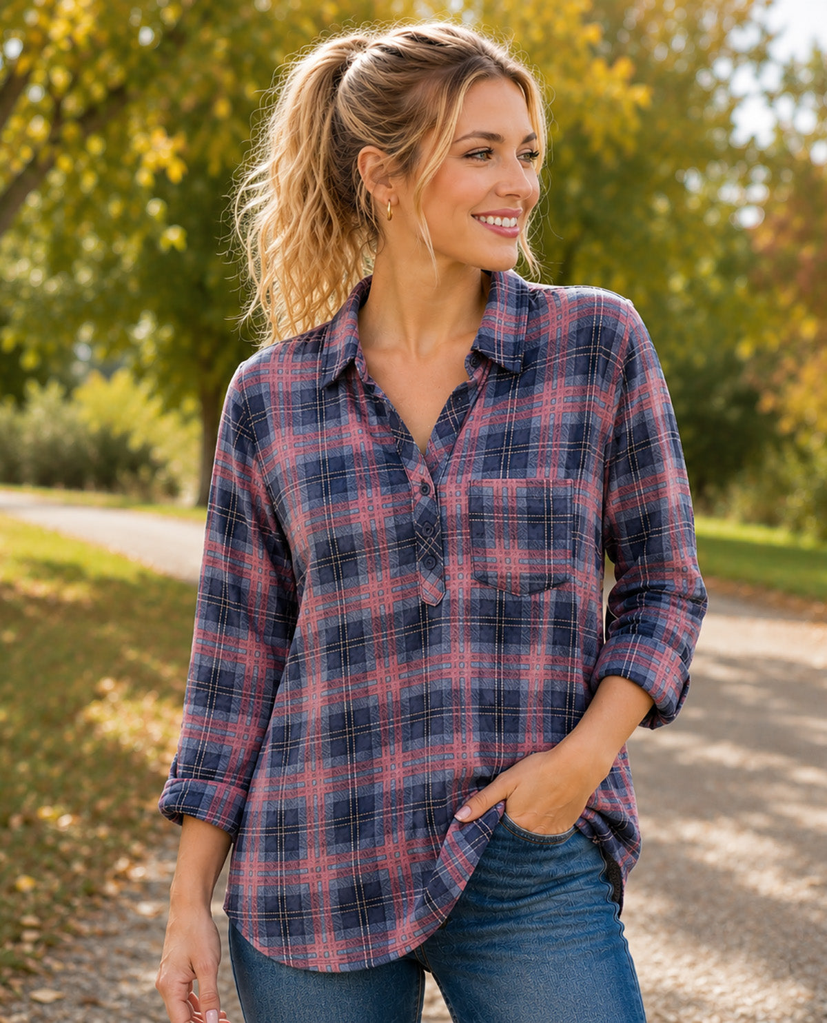 A woman wearing a long-sleeve plaid button-up shirt in navy and dusty pink, paired with blue jeans, standing outdoors on a path with trees in the background.