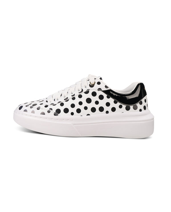 A white low-top sneaker featuring a black polka dot pattern across the upper, white laces, and a thick white platform sole with a black heel accent.