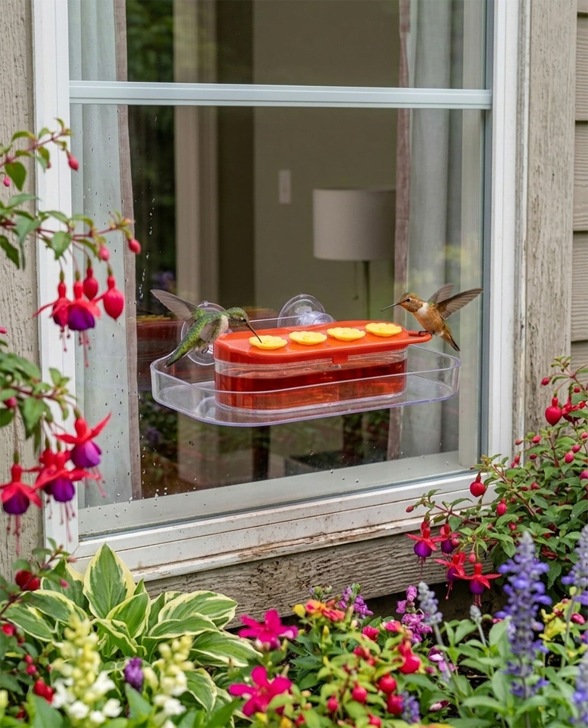 A clear plastic hummingbird feeder with orange-red wax melts sits on a window ledge, with two hummingbirds feeding. The window is framed by lush green plants and colorful flowers.