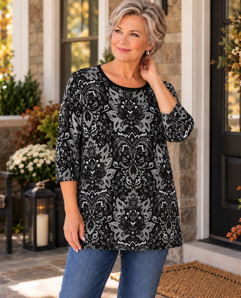 A woman wearing a black and gray patterned three-quarter sleeve tunic top with blue jeans, standing on a stone porch.