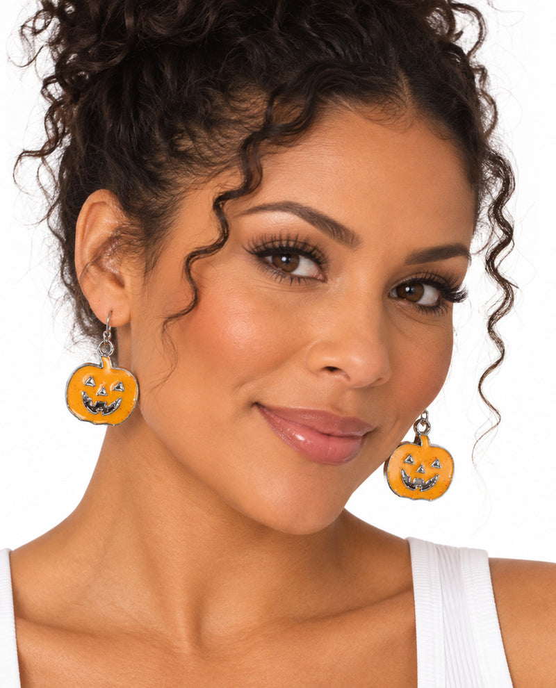 A close-up of a woman wearing orange jack-o'-lantern dangle earrings with black facial details and silver hooks, set against a plain white background.