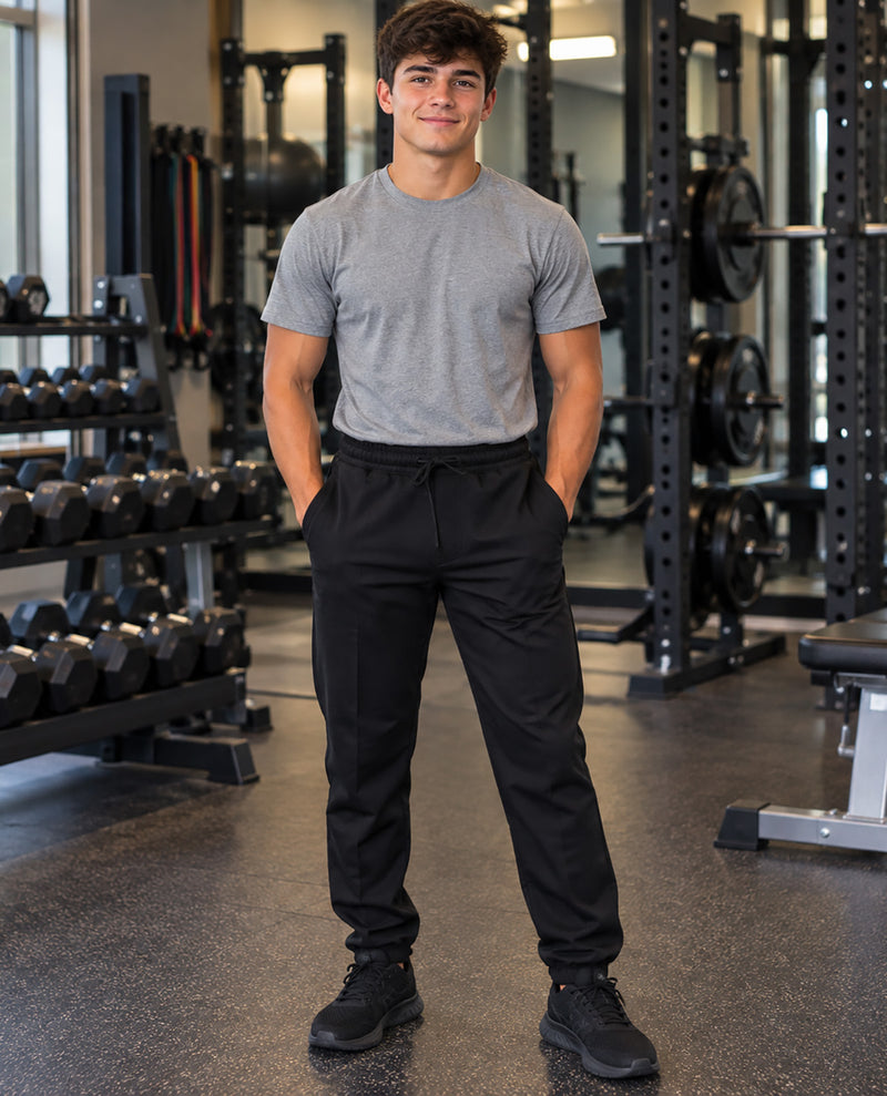 A man standing in a gym wearing a heather gray crewneck t-shirt and black drawstring sweatpants, with weight racks and dumbbells visible in the background.