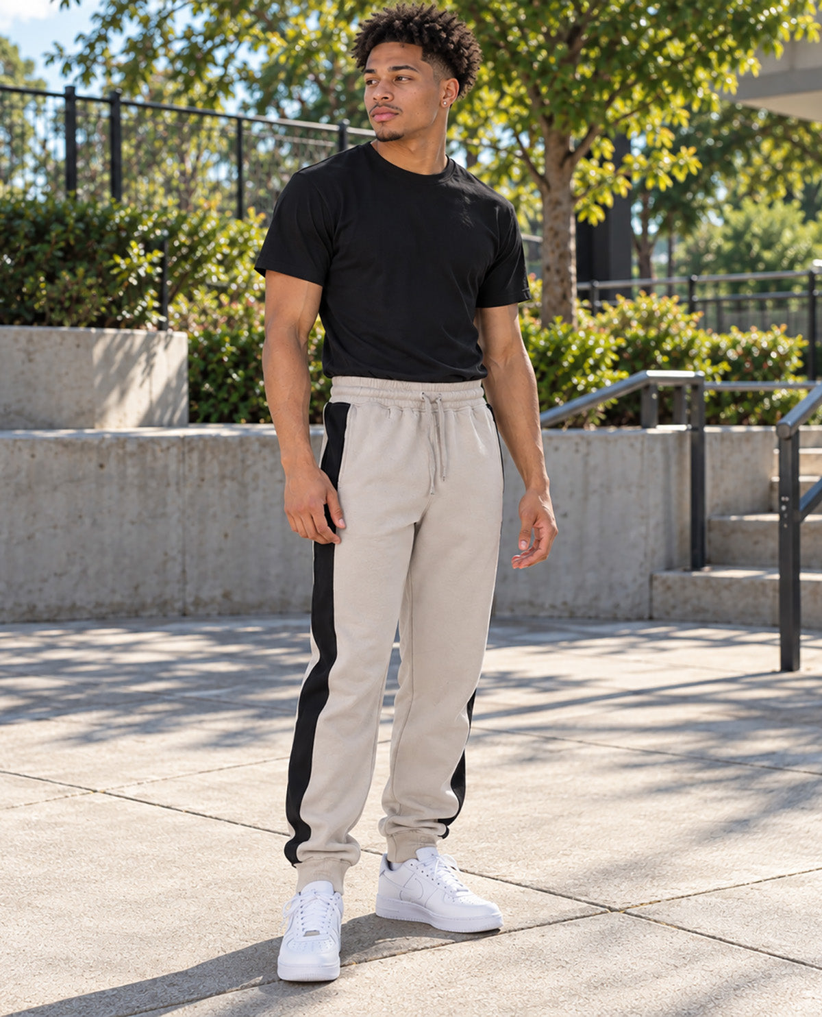 A man wearing beige drawstring sweatpants with black side panels, a black t-shirt, and white sneakers, standing outdoors on a concrete walkway with trees and a fence in the background.
