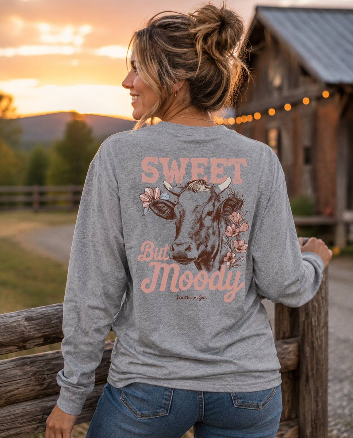 Woman wearing a gray long-sleeve shirt with a cow graphic and 'Sweet But Moody' text at sunset.